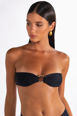 A woman wearing a black bandeau bikini top with vintage gold ring hardware at the front and an adjustable tie at the back.